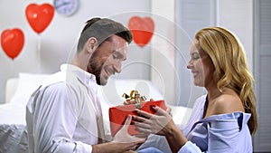 Seductive young female giving red gift box to surprised boyfriend, anniversary