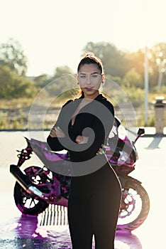 Seductive young woman in tight fitting black suit poses near sport motorcycle at self service car wash.