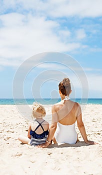 Seen from behind mother and child in swimsuits sitting at beach