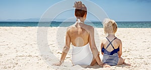 Seen from behind mother and child in swimsuits sitting at beach