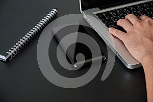 Selected focus hand using laptop on black desk office  with smartphone and spiral notebook