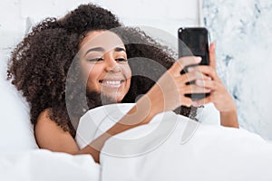 Selective focus of african american young woman using smartphone in bed during morning time