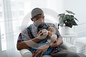 Selective focus on African father hand holding milk bottle and feeding milk to cute toddler baby lying in his arms at home