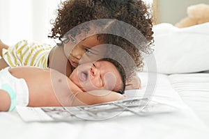 Selective focus of African little older sister trying to comforting newborn baby brother crying lying on white bed at home