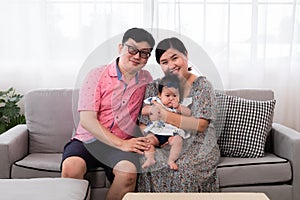 Selective focus Asian happy family sitting on cozy sofa cuddling with adorable infant take a photo in living room. Newborn baby