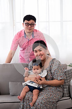 Selective focus Asian mother holding adorable infant sitting on cozy sofa take a photo with father in living room. Newborn baby