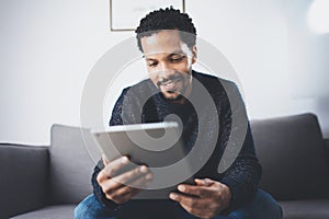 Selective focus.Attractive bearded African man reading news on digital tablet while sitting sofa in his modern studio