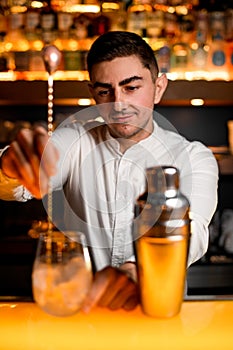 Selective focus of bartender stirring alcoholic cocktail with bar spoon on yellow bar surface