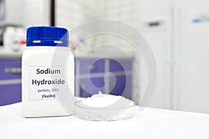 Selective focus of a bottle of pure sodium hydroxide or NaOH chemical compound beside a petri dish with white solid pellets.