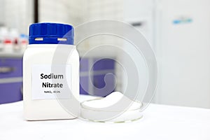 Selective focus of a bottle of sodium nitrate preservative compound beside a petri dish with white crystalline powder.