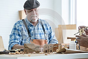 Selective focus on car and house wooden toy on table. Caucasian senior old white bearded man carpenter in apron and hat working in
