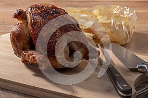 Select focus of a spit-roasted chicken on a wooden board with an out of focus bag of potato chips
