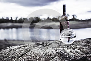 Selective focus of a crystal ball on a rock reflecting a bottle of wine and a beautiful lake behind