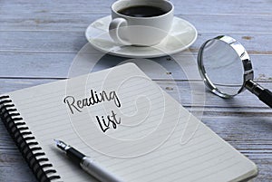 Selective focus of a cup of coffee,magnifying glass,pen and notebook written with Reading List on wooden background