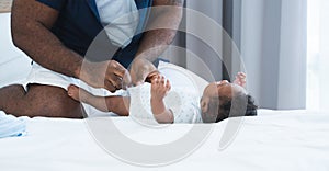 Selective focus on dad hands. Father changing diaper and clothes for African Nigerian newborn baby lying on white bed in bedroom.