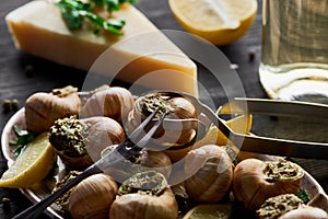 Selective focus of delicious cooked escargots with lemon, parmesan, cutlery, parsley and white wine on black wooden