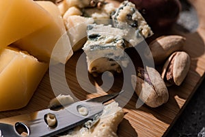 Selective focus of dorblu pressed by knife next to pieces of grana padano and pistachios on cutting board.