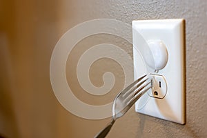 Selective focus on edge of a metal fork about to be inserted into an open electrical outlet socket. The top outlet has a plastic