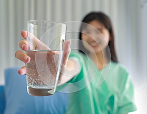 Selective focus on glass. Young inpatient woman  who is getting well from her ailment smiling on to a camera and holding a glass