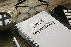 Selective focus of a glasses, coffee,mobile phone,pen and notebook written with Roles and Responsibilities on wooden background