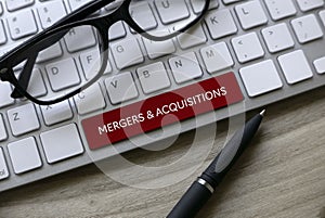 Selective focus of glasses, pen and computer keyboard with red key written with Mergers and Acquisitions on a wooden background