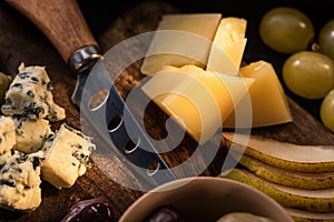 Selective focus of grana padano with bowl of olives, knife, slices of pear, grapes and dorblu on cutting board.