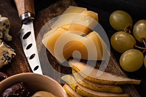Selective focus of grana padano with pieces of pear, bowl of olives, knife, dorblu, grapes on cutting board.