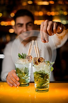 selective focus on hand of bartender gently pouring sugar on glass with mojito on the bar