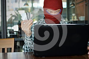 Selective focus on hands of Masked hacker wearing a balaclava holding credit card between stealing data from laptop. Internet crim
