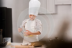 selective focus of happy boy in chef hat and apron whisking eggs in bowl at table