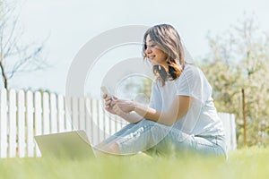 Focus of happy woman using smartphone near laptop while sitting on grass