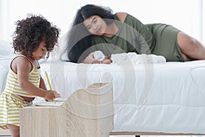 Selective focus of little African girl playing drawing book while mother looking at her kid and also feeding milk from bottle to i