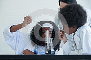 Selective focus on Little African girl`s hand mixing color liquid by dropper in flasks with scientist boy and Asian man teacher.