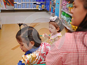 Little Asian baby girl  furthest  looking and standing in a shopping cart with her sister while her mother photo