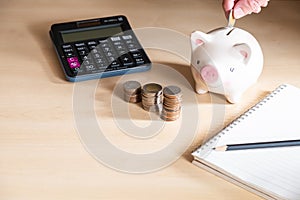 Selective focus at men hand put coin into piggy bank. With blurred calculator and increase stack of money on wooden desk. Personal