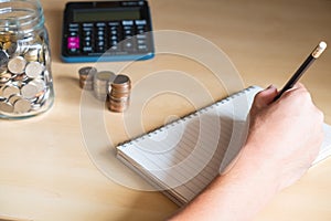 Selective focus at men hand while writing personal saving plan on the paper with blurred stack of money coins, glass jar coin and