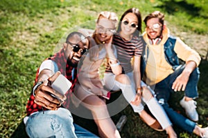 selective focus of multiethnic friends taking selfie on smartphone while resting on green lawn