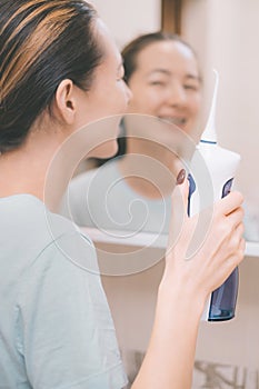 Selective focus on oral irrigator in hand. A young woman smiles and looks in the mirror in the background.