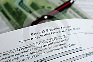 Selective focus photo of paycheck protection program borrower application form revised, on a background of dollar bills and a pen