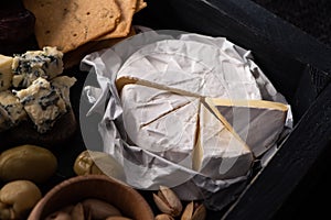 Selective focus of pieces of camembert next to dorblu, dried olives, crackers and pistachios on tray on black background