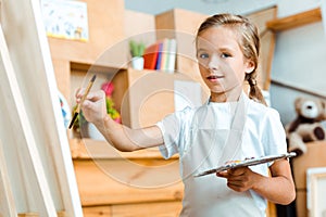 selective focus of positive kid holding palette and paintbrush.