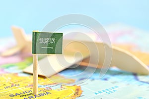 Selective focus of Saudi Arabia flag in blurry world map and airplane model. Travel and tourism destination concept.