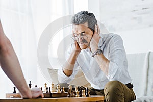 Selective focus of senior man with mustache covering ears while playing chess with friend