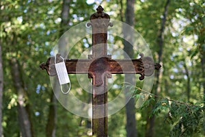Selective focus shot of a rusty metal cross with a facemask hanging on it in an old cemetery