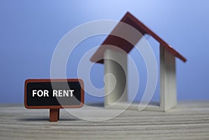 Selective focus of small wooden house and red wooden tag written with For Rent on wooden table with a blue background