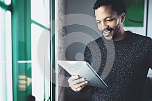 Selective focus.Smiling bearded African man reading book on digital tablet while standing near the window in his modern