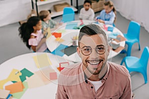selective focus of smiling teacher in eyeglasses and interracial kids at table