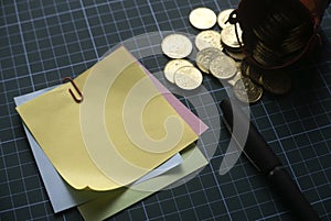 Selective focus of stack of memo note with red bucket of gold coins and pen on green square background.Copy space for text, logo