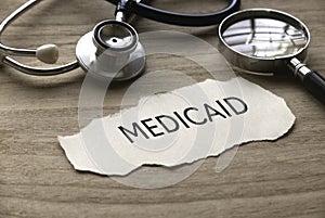 Selective focus of stethoscope, magnifying glass and piece of paper written with Medicaid on wooden background