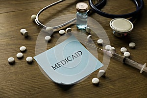 Selective focus of stethoscope, white medical pills, syringe and blue memo note written with Medicaid on wooden background